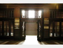 Holzvertäfelter Eingangsbereich eines historischen Gebäudes mit einer zentralen Treppe, die zu einer hell beleuchteten oberen Etage führt. Rechts und links der geöffneten Glastüren befinden sich Übersichtstafeln mit Beschriftungen.