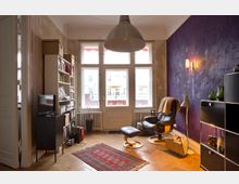 Wohnzimmer mit Parkettboden und großen Fenstern, die viel Tageslicht hereinlassen. Eine Wandecke ist in Lila gestrichen, daneben stehen ein schwarzer Sessel mit Hocker, ein Regal voller Bücher, ein TV-Schrank und ein Sideboard. Ein Teppich und eine hängende Metalllampe ergänzen die Einrichtung.