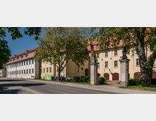 Historisches Gebäudeensemble mit hellen Fassaden und rotem Ziegeldach, flankiert von großen Bäumen. Ein Tor mit steinernen Säulen führt in einen Innenhof, daneben befindet sich eine Straße mit Gehweg.