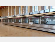 Weitläufige Abfertigungshalle eines Flughafens mit einer Gepäckförderanlage im Vordergrund, darüber eine Reihe von geschlossenen Schaltern mit Glasabtrennungen und roten Schildern, im Hintergrund hohe Fenster sowie beige Steinwände.