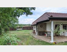 Ein Garten mit einem kleinen Gebäude mit rotem Ziegeldach und einer überdachten Terrasse. Die Terrasse hat eine Sitzbank, einen Tisch und eine Dartscheibe an der Steinwand; im Hintergrund sind ein Holzzaun, ein Tor und Felder zu sehen, umgeben von grünen Bäumen.
