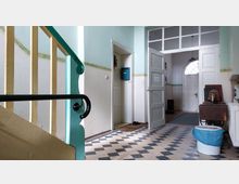 Treppenhaus eines Altbaus mit mintgrünen Wänden und gelbem Treppengeländer. Der Boden ist mit schwarz-weiß karierten Fliesen belegt, und mehrere offene Türen führen in angrenzende Räume; im Hintergrund ist eine weiße Holztür mit rundem Fenster zu sehen.