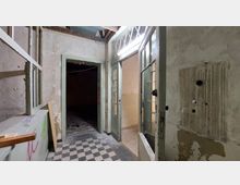 Ein verlassener, heruntergekommener Innenraum mit abblätternder Farbe an den Wänden, einem schwarz-weiß karierten Boden und einfachen, verglasten Doppeltüren. Die Decke zeigt freiliegende Balken, und der Raum dahinter wirkt dunkel und leer.