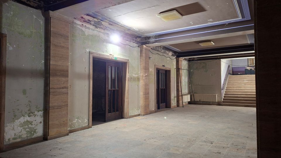 Verlassene Innenhalle eines älteren Gebäudes mit abblätternder Wandfarbe, Steinverkleidungen an Pfeilern und zwei dunklen Holztüren. Rechts führt eine breite Treppe nach oben; an der Decke sind rechteckige Leuchten und freiliegende Kabel zu sehen.