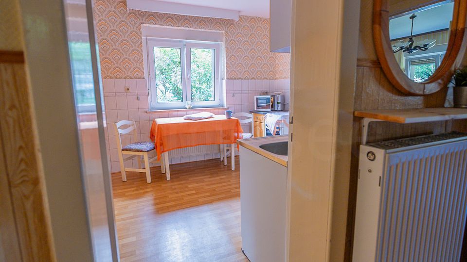 Blick in eine kleine Wohnküche mit Holzfußboden, gemusterter Tapete und einem Fenster über einem Tisch mit orangefarbener Tischdecke und zwei Stühlen. Rechts sind Küchenzeile mit Spüle sowie ein Regal mit Spiegel und einem Heizkörper zu sehen.