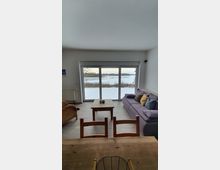 Wohnzimmer mit großen Glas-Schiebetüren, die auf eine verschneite Landschaft mit einem See im Hintergrund blicken. Im Raum stehen ein Holztisch, eine lila Couch mit Kissen, Holzstühle und ein Rattansessel.