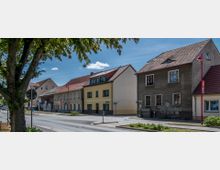 Straßenszene in einer Kleinstadt mit Wohn- und Geschäftshäusern im traditionellen Baustil, erkennbar an Satteldächern und verputzten Fassaden. Im Vordergrund steht ein Baum, und am Straßenrand sind Verkehrsschilder sowie eine laternenartige rote Straßenleuchte sichtbar.