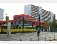Straßenszene mit einer gelben Straßenbahn im Vordergrund, einer roten, modernen Gebäudefassade in der Mitte und einem großen Wohnkomplex mit Balkonen im Hintergrund. Ampeln, Passanten und Haltestellenzeichen betonen den städtischen Charakter.