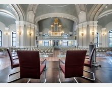 Innenraum einer historischen Kirche mit weißer Stuckverzierung und großen Rundbogenfenstern. Im Vordergrund stehen zwei rote Sessel und ein Pult, im Hintergrund sind Stuhlreihen, eine Empore und eine vergoldete Orgel sichtbar.