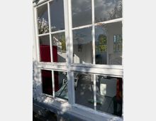 Eine Nahaufnahme eines Gebäudefensters mit weiß gestrichenem Holzrahmen, der bereits abgenutzt ist. Durch die Glasscheiben ist ein heller Innenraum mit Holzboden, einem Besen und Möbeln sichtbar; draußen spiegelt sich ein Baum im Glas.