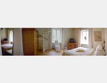 Helles Schlafzimmer mit Holzfußboden, einem weißen Metallbett, einem Kleiderschrank aus Holz und einer Wendeltreppe daneben. Zwei Fenster mit weißen Vorhängen lassen Tageslicht herein, und ein kleiner Fernseher steht auf einer Holzkommode.