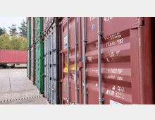 Reihe von Frachtcontainern in verschiedenen Farben, hauptsächlich rot, grün und grau, aufgereiht auf einem Betonplatz; im Hintergrund ein weiterer Container auf einem Eisenbahnwaggon und Bäume.