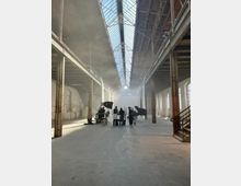 Große, leere Industriehalle mit hohen Decken, sichtbarer Stahlkonstruktion und Oberlichtern, durch die Sonnenlicht einfällt. Im Zentrum des Raumes befindet sich eine Gruppe von Personen umgeben von filmischem Equipment und einem Flügel, was auf eine Aufnahme- oder Filmproduktion hinweist.