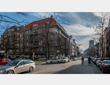 Straßenansicht mit mehrstöckigen Wohngebäuden im Altbaustil, roten Dächern und großen Fenstern, flankiert von parkenden Autos auf beiden Straßenseiten. Im Hintergrund ist eine Kirche mit Turm zu sehen, und kahle Bäume säumen die Straße.