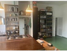 Raum mit Küchen- und Büroelementen: Im Vordergrund eine Holzarbeitsfläche mit eingebautem Gasherd, im Hintergrund offene Regale mit Küchenutensilien, Bücher und Büroordnern. Grüne Hängelampen und ein grüner Boden ergänzen die Einrichtung, links sind Waschbecken sichtbar und rechts eine Tür.