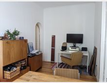 Ein schlicht eingerichtetes Zimmer mit weiß gestrichenen Wänden und Holzboden. Links steht ein Holzschrank mit darauf platzierten Pflanzen und Büchern, rechts ein Schreibtisch mit einem Computer, Lautsprechern und einer Musikanlage, davor ein gepolsterter Stuhl.