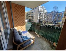 Ein Balkon mit einem Holzsofa und Kissen, umgeben von orangefarbenen Wänden. Im Hintergrund sind moderne Wohngebäude mit Balkonen sowie ein grüner Sichtschutz und eine Lichterkette zu sehen.
