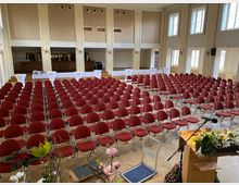 Großer Versammlungsraum mit Reihen von roten Stühlen auf einem hellen Holzboden, einer Bühne mit Blumenarrangement und Mikrofon im Vordergrund. Im Hintergrund befinden sich Stehtische sowie Fenster mit hellem Tageslicht, und die Raumgestaltung ist durch beige Wände und hohe Decken geprägt.