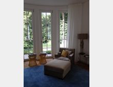 Wohnzimmer mit großen bodentiefen Fenstern, die Blick auf einen grünen Garten bieten. Im Raum befinden sich eine graue Liege mit gelbem Kissen, zwei kleine Beistelltische aus Rattan, eine Stehlampe und ein blauer Teppich.