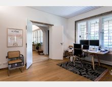 Helles Büro mit Holzboden, einem Schreibtisch mit zwei Monitoren vor einer Fensterfront mit verzierten Glaselementen. Im Vordergrund steht ein Flechtstuhl, daneben führt eine geöffnete Tür in einen angrenzenden Raum mit Pflanzen und dekorativen Elementen.