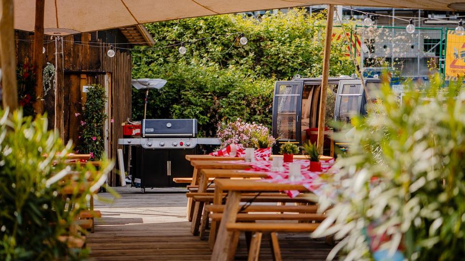 Überdachter Außenbereich mit Holzterrasse und langen Picknicktischen mit rot-weiß karierten Tischdecken und Pflanzendekoration. Im Hintergrund stehen ein großer Gasgrill neben einer Holzwand und eine kleine, verglaste Kabine; Lichterketten und dichtes Grün umgeben den Bereich.
