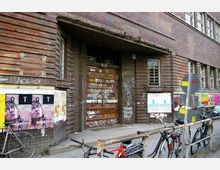 Backsteinfassade eines Gebäudes mit einer Holztür, die mit Graffiti bedeckt ist. Im Vordergrund stehen Fahrräder an einem Metallgeländer, und auf der Wand sind mehrere Plakate und Aufkleber angebracht.