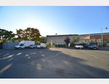 Parkplatz mit mehreren geparkten Fahrzeugen, darunter Lieferwagen und Autos, vor einer flachen, modernen Gebäudefassade mit großen Fenstern und Klinker-Elementen. Im Hintergrund stehen Bäume, teils in Herbstfarben, unter klarem Himmel.