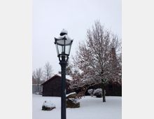 Eine verschneite Szene mit einer schwarzen Straßenlaterne im Vordergrund, die von Schnee bedeckt ist. Im Hintergrund steht ein Gebäude mit einem roten Ziegeldach, umgeben von schneebedeckten Bäumen und Boden.