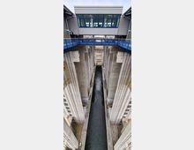 Blick von oben auf eine moderne Schiffshebewerksanlage mit hohen Betonwänden, einem schmalen Wasserkanal in der Mitte und einem verglasten Kontrollraum oben quer über der Anlage. Geländer und technische Elemente sind entlang der Struktur sichtbar.