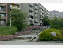 Mehrstöckige Wohnhäuser aus Beton mit Balkonen in Reihenanordnung, flankiert von Grünflächen und Bäumen. Im Vordergrund sind mehrere gestufte Treppen mit Geländern und umliegenden Sträuchern zu sehen.