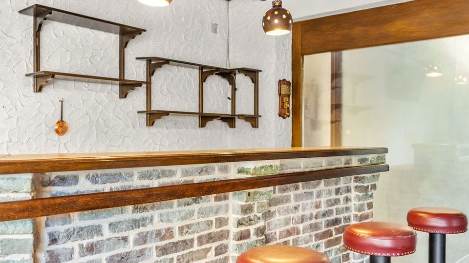 Innenraum einer kleinen Bar mit gemauerter Theke, Holzbalkendecke und zwei hängenden Kugelleuchten. Vor der Theke stehen mehrere hohe Barhocker mit roten Lederpolstern; an der weißen Wand sind offene Regale und rechts eine Milchglasschiebetür.