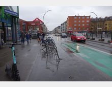 Straßenszene in einer belebten städtischen Gegend, mit einem Supermarkt (Rewe) und anderen Gebäuden im Hintergrund. Im Vordergrund stehen Fahrräder und ein E-Scooter, daneben verläuft ein nasser Radweg, während Autos auf der Straße fahren.