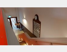 Treppenhaus in einem öffentlichen Gebäude mit einer alten hölzernen Anzeigetafel und einer Uhr an der Wand. Neben der Treppe führt eine Tür mit Glaselementen ins Freie, und die Wände sind teilweise gefliest.