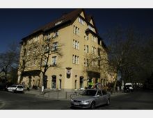 Eckgebäude in gelber Putzfassade mit mehreren Stockwerken und Satteldach, umgeben von kahlen Bäumen. Im Erdgeschoss befinden sich Schaufenster und Geschäfte, während Autos und Passanten die Straße beleben.