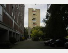 Ein schmaler Hof zwischen zwei Gebäuden: links ein mehrstöckiger Bau mit roten Ziegeln und vielen Fenstern, rechts Bäume und parkende Autos. Im Hintergrund steht ein markanter, grauer Hochhausturm mit Sichtbetonfassade und mehreren Balkonen.