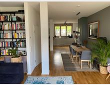 Ein modernes Wohnzimmer mit Holzboden, einem offenen Regal voller Bücher an der linken Wand und einem Essbereich mit Holztisch und grauen Stühlen rechts. Im Hintergrund eine offene Küche mit weißen Schränken und Fenstern, die Tageslicht hereinlassen.