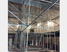 Metallgerüstkonstruktion in einem großen Innenraum mit Steinwänden und mehreren Wandleuchten. Die Decke ist unverkleidet, und der Raum scheint sich in einer Um- oder Neubauphase zu befinden.