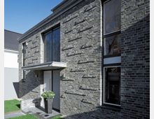 Moderne Wohnhausfassade aus dunklem Ziegelstein, mit großen Fenstern und einem rechteckigen Vordach über der Eingangstür. Vor der Tür steht ein hoher schwarzer Pflanztopf mit grünem Gewächs, und der angrenzende Garten zeigt einen schmalen Streifen Rasen.