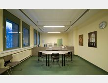 Ein einfacher Besprechungsraum mit einer gelb gestrichenen Wand, vier großen Fenstern, einem Heizungskörper und grünem Bodenbelag. In der Mitte steht ein trapezförmiger Tisch mit vier Stühlen, dahinter ein niedriger Tresen mit Bürostühlen; an der Wand hängen zwei Bilder und eine Wanduhr.