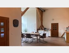 Ein großzügiger Raum mit Holzbalken an der Decke, einem Esstisch mit sechs Stühlen, einer Küchenzeile an der Rückwand und einer Holztür. Links an der Wand hängt eine Dartscheibe, und eine Zimmerpflanze steht in der Ecke vor hellen Wänden.