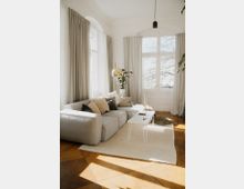 Helles Wohnzimmer mit einer grauen Eckcouch, dekoriert mit Kissen, einem kleinen weißen Couchtisch und einer goldenen Stehleuchte. Große Fenster mit beigen Vorhängen lassen viel Tageslicht herein; der Boden ist aus Holz und mit einem weißen Teppich bedeckt.