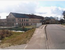 Großes, historisches Fabrikgebäude aus Backstein mit vielen Fenstern und einem Satteldach, daneben eine asphaltierte Straße mit Gehweg. Im Vordergrund befindet sich eine Rasenfläche und ein kleiner Wasserkanal mit Geländer.