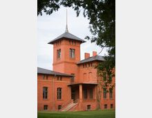 Backsteingebäude mit einem hohen Turm, spitzem Dach und mehreren großen Fenstern, umgeben von einem grünen Rasen und teils eingerahmt von Baumzweigen im Vordergrund.