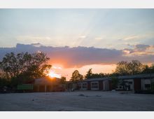 Ein Industriehof bei Sonnenuntergang, mit mehreren flachen, roten Gebäuden und einem Baumhain im Hintergrund. Die Sonne scheint hinter einer großen Wolke hervor, wodurch einige Sonnenstrahlen sichtbar werden.