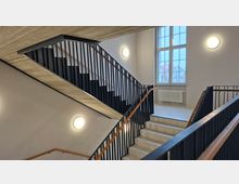 Innenansicht eines Treppenhauses mit mehreren Betonläufen, schwarzen Metallgeländern und Holzhandläufen. Helle Wände mit runden Wandleuchten sowie ein großes Sprossenfenster mit Heizkörper darunter prägen den Raum.