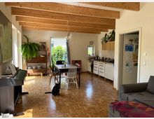 Wohnzimmer mit offener Küche und Holzdecke. Im Raum stehen ein Esstisch mit Stühlen, ein Sofa und ein Klavier; Pflanzen und Regale schmücken den Bereich, und durch die Fenster fällt Tageslicht auf den Parkettboden.