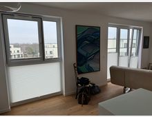 Helles Wohnzimmer mit Holzboden, zwei großen Fenstern mit halb geschlossenen Plissees und Blick auf moderne Wohngebäude. An der Wand hängt ein abstraktes Gemälde, ein weißes Sofa steht im Raum, und Taschen sowie ein Stuhl befinden sich am Fenster.