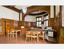 Innenraum mit Holzvertäfelung im Jugendstil, ausgestattet mit Schreibtischen, Stühlen und moderner Büroausstattung wie Computer und Lampen. Hohe Fenster mit Bleiglas und ein geschwungener Holzbogen betonen die historische Architektur.