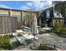 Ein Außenbereich eines Cafés mit gepflastertem Boden, weißen Tischen, grünen Stühlen mit Polstern und geschlossenen Sonnenschirmen. Im Hintergrund ist ein Fachwerkhaus mit dunklem Schieferdach und kleinen Dachfenstern sowie eine Glaswand zu sehen, umgeben von Bäumen und Pflanzen.