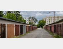 Eine schmale Gasse mit Reihen von Garagen auf beiden Seiten, deren Tore in verschiedenen Farben gestrichen sind. Im Hintergrund sind Wohngebäude und Bäume zu sehen, unter bewölktem Himmel.